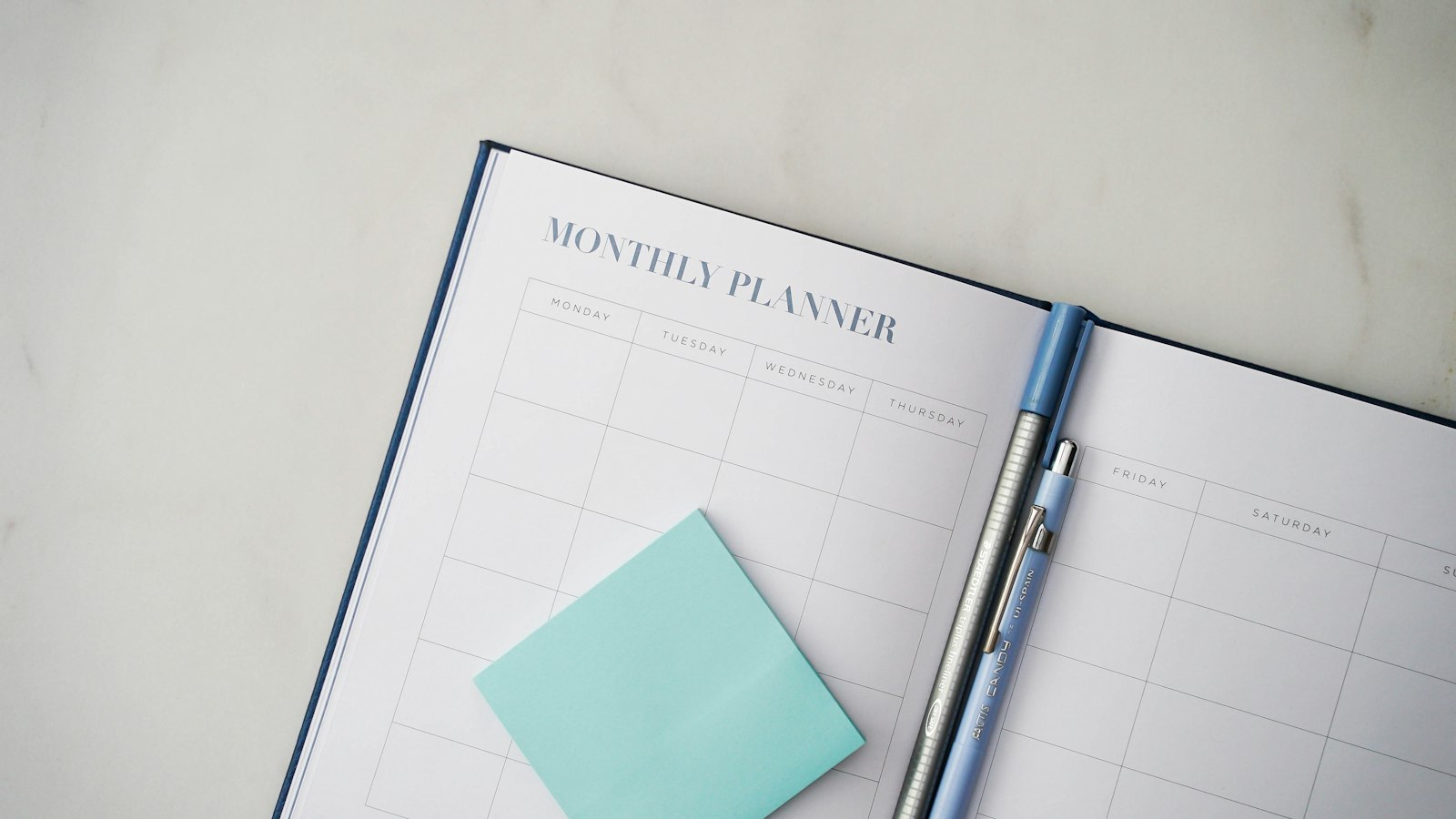 An open weekly planner with a pen resting across its pages on a marble desk.