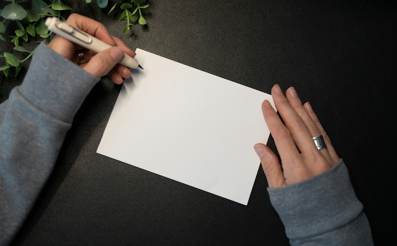 Hands writing a note on a blank card, with a sprig of greenery on the desk.