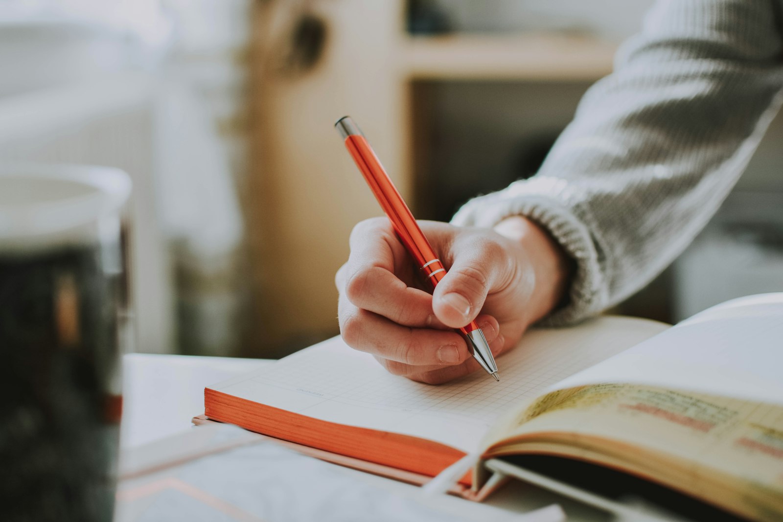 A hand holding a red pen, writing in an open notebook beside a cup.